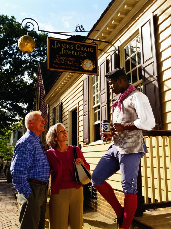 A man dressed in Colonial clothing stands outside of his shop talking to two older people