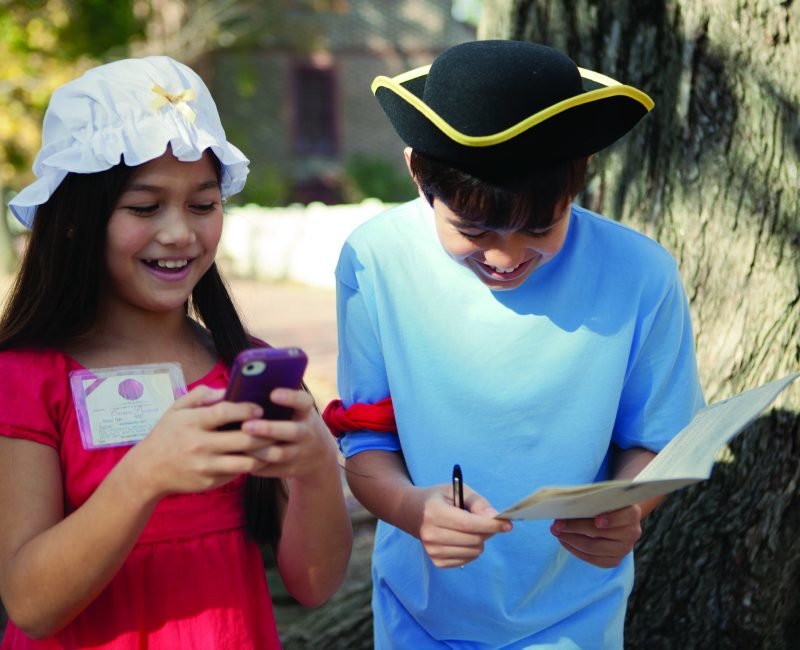 Two children playing a scavenger hunt game with the girl on her phone and the boy writing things dow