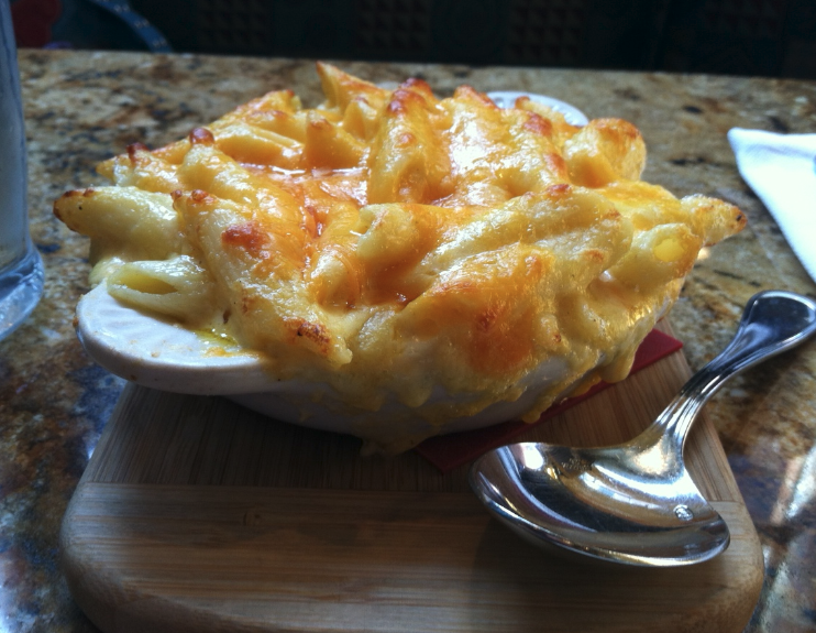 A bowl of mac and cheese on a wooden serving board with a large silver spoon