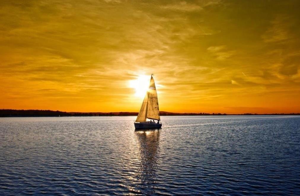 A sailboat on the water at sunset with a deep yellow and orange sky behind the boat